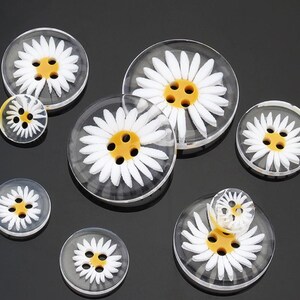 May include: Assortment of clear plastic buttons with white daisy designs and yellow centres.