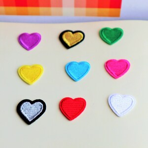 May include: Nine felt heart patches in various colors, including red, yellow, blue, green, pink, white, black, and gold. The patches are arranged in a grid pattern on a white surface.