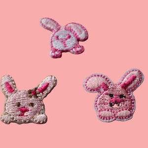 May include: Three embroidered bunny patches in pink and white, each with a unique pose. The bunnies have pink ears, noses, and feet, with white accents. The background is a solid light pink.