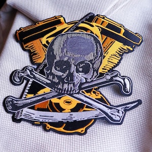 May include: A black and white skull and crossbones patch with a yellow engine block behind it. The patch is on a white fabric background.