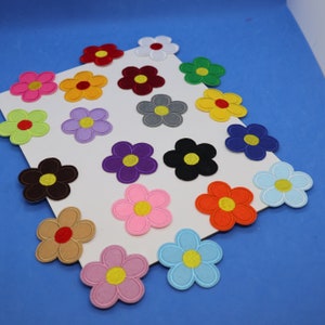 May include: Assortment of 16 colourful felt flower patches with yellow centres. The patches are arranged in rows on a white background.
