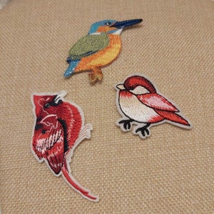 May include: Three embroidered bird patches: a kingfisher with blue, green, and orange colours, a red cardinal, and a small red and white bird. These patches are suitable for adding to clothing or accessories.