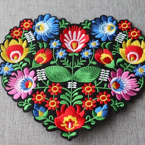 May include: A colourful embroidered heart-shaped patch with a floral design. The patch features red, yellow, blue, green, pink and white flowers on a black background.