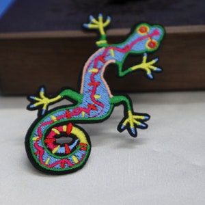 May include: A colourful embroidered patch of a green, blue and pink lizard with a curled tail. The lizard has yellow feet and red eyes.