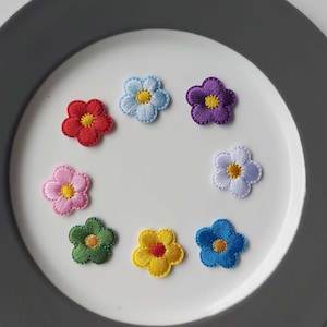 May include: Assortment of eight embroidered flower patches in various colours, including red, yellow, blue, green, purple, and white. The patches are all the same size and shape, and they are arranged in a circle on a white surface.