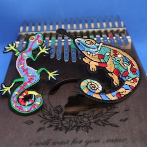 May include: A wooden musical instrument with a colourful embroidered lizard and chameleon design. The instrument has a phrase "I will wait for you, my love" engraved on the surface.