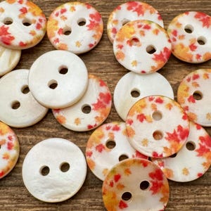 May include: A collection of round, two-hole buttons featuring a mix of solid white pearlised surfaces and white surfaces printed with small red, orange, and yellow autumn maple leaf patterns. Each button has a smooth, circular design.