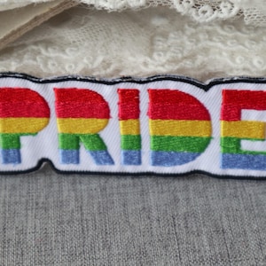 Peut inclure: Écusson brodé avec le mot "PRIDE" en couleurs arc-en-ciel : rouge, orange, jaune, vert, bleu et violet. L'écusson a un fond blanc et une bordure noire. Il est de forme rectangulaire et légèrement incurvé.