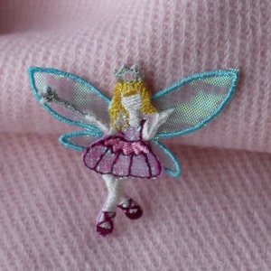 May include: A pink and blue fairy with a crown and wings, embroidered on a pink fabric.