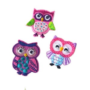 May include: Three embroidered owl patches in pink, purple, and blue. Each owl has large, round eyes and is embellished with colourful stitching. These patches are suitable for decorating clothing or accessories.