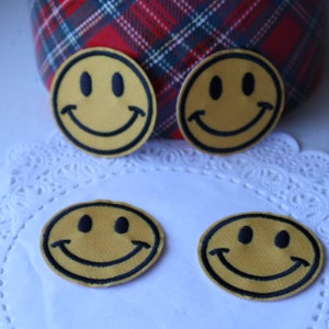 May include: Four yellow smiley face patches with black outlines and features. The patches are arranged on a white lace surface, with a plaid fabric in the background. The patches are round and have a classic smiley face design.