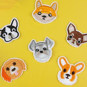 May include: Six embroidered iron-on patches featuring different dog breeds. The patches are white with colourful embroidered details. The dogs include a chihuahua, a schnauzer, a corgi, a golden retriever, a fox, and a french bulldog.