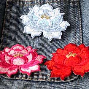 May include: Embroidered lotus flower patches in light blue, pink, and red. The patches feature detailed stitching and a gold centre, set against a denim background. These decorative patches can be used to embellish clothing or accessories.