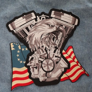 May include: A black and white embroidered patch featuring a stylized eagle head with a motorcycle engine behind it. The eagle is looking to the right and the engine is partially obscured by the eagle's head. The patch is surrounded by two American flags, one on each side.