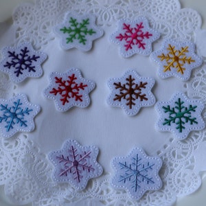 May include: Assortment of ten snowflake-shaped felt patches in various colors, including blue, green, red, pink, brown, and white. The patches are arranged on a white doily.