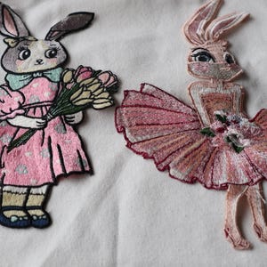 Peut inclure: Deux patchs brodés de lapins roses. Un lapin porte une robe rose et tient un bouquet de fleurs. L'autre lapin porte un tutu rose et tient une seule fleur.