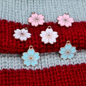 May include: Six metal flower charms in pink, white, and blue. The charms are in the shape of a five-petal flower with a gold center. The charms are on a striped knitted fabric.