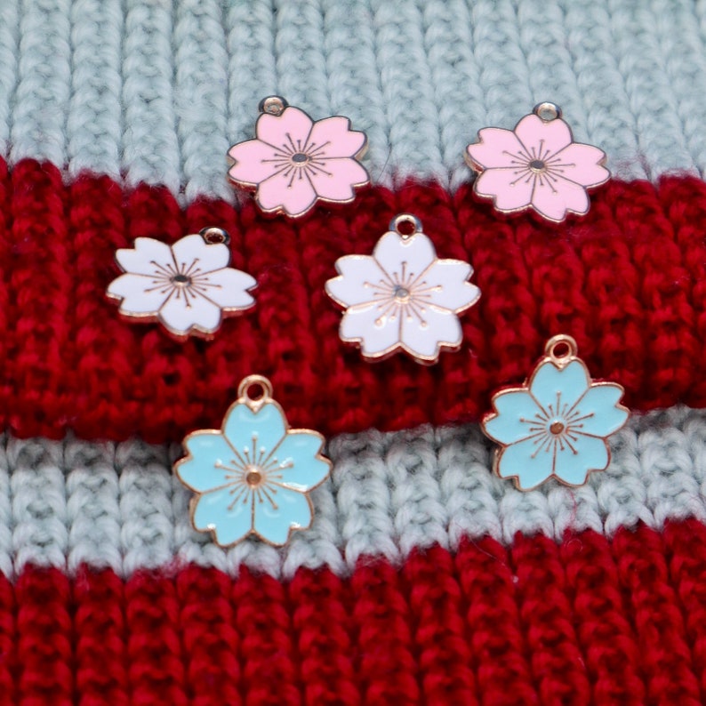 May include: Six metal flower charms in pink, white, and blue. The charms are in the shape of a five-petal flower with a gold center. The charms are on a striped knitted fabric.