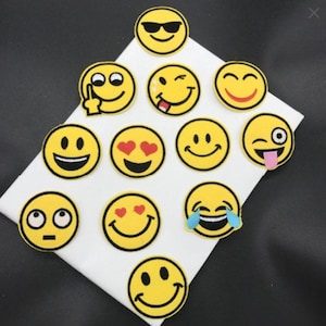 Peut inclure: Plusieurs écussons emoji jaunes avec diverses expressions, notamment des lunettes de soleil, des cœurs à la place des yeux et un clin d'œil. Les écussons sont ronds, bordés de noir et disposés sur une surface blanche.