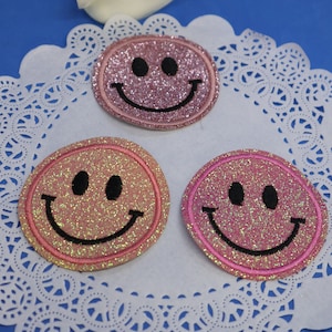 Peut inclure: Trois patchs thermocollants en forme de smiley en paillettes roses. Les patchs sont ronds et ont un contour noir. Ils sont sur une dentelle blanche.