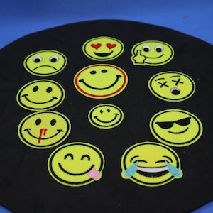 Peut inclure: Un ensemble de patchs smiley jaunes avec diverses expressions, notamment souriantes, clignotantes et pleurantes. Les patchs sont brodés avec des contours et des détails noirs, sur un fond noir.
