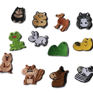 May include: A collection of twelve embroidered fabric patches featuring various animals and shapes, including a black bear, a tan hamster, a brown camel, a gray hamster, a green frog, a gray rabbit, a green mountain, a yellow shape, a quokka, a squirrel, a horse head, and a zebra.