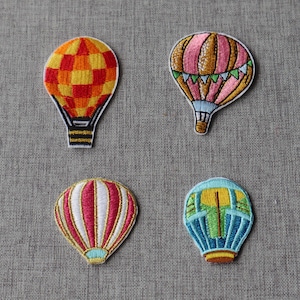 May include: Four embroidered hot air balloon patches. The top left is orange and yellow checkered. The top right is pink and brown with green bunting. The bottom left has vertical pink and white stripes. The bottom right is blue and yellow.