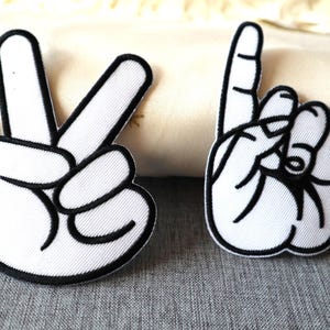May include: Two embroidered patches featuring hand gestures. One patch shows the peace sign with white fabric and a black outline. The other patch depicts the 'rock on' hand gesture, also with white fabric and a black outline.