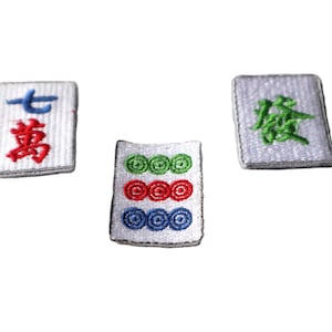 Peut inclure: Trois tuiles de mahjong blanches avec des caractères et des symboles chinois brodés. La première tuile porte les caractères chinois "七萬" (sept de dix mille), la deuxième tuile porte trois rangées de cercles en vert, rouge et bleu, et la troisième tuile porte le caractère chinois "發" (prospérité).