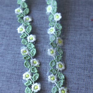 May include: Two strips of floral lace. The lace features white daisy-like flowers with yellow centres and green leaves, set against a light green background. The lace is likely used for embellishing crafts or clothing.