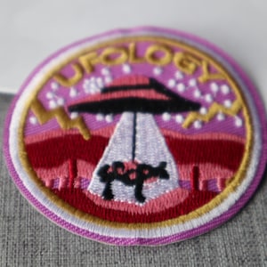 May include: Embroidered patch with a circular design featuring a UFO and the word "UFOLOGY" in gold lettering. The patch has a pink and purple background with a red and white border. The UFO is black and white.