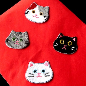 May include: Four embroidered cat patches in various colors: white, gray, and black. Each patch features a different cat face design with embroidered details like eyes, whiskers, and a pink mouth. The patches are set against a bright red background.