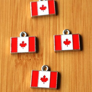 May include: Four silver charms featuring the Canadian flag. Each charm is a small rectangular shape with a red and white Canadian flag design. The charms have a silver loop at the top for attaching to a necklace or bracelet.