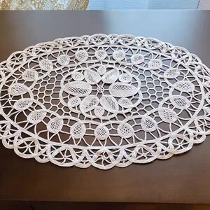 May include: Oval white lace doily with a detailed floral and geometric pattern. The doily is made of a delicate, open-weave fabric and is placed on a dark brown surface. The edges are scalloped.