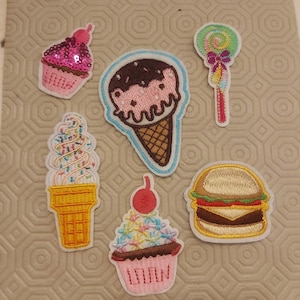 May include: Embroidered patches of food items. Includes a pink sequined cupcake, ice cream cone, lollipop, soft serve ice cream, cupcake with flowers, and a hamburger. Each patch has a white border.