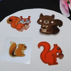 May include: Four embroidered iron-on patches featuring squirrels. Two of the patches are orange, one is brown, and one is a multi-coloured orange and brown. Each patch features a squirrel holding a nut.