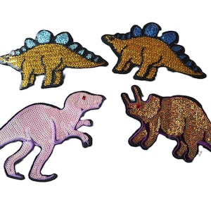 May include: Four iron-on patches featuring different dinosaurs. The patches are made of sequins and have a shiny finish. The dinosaurs are a pink T-Rex, a gold stegosaurus, a gold triceratops, and a gold brachiosaurus.