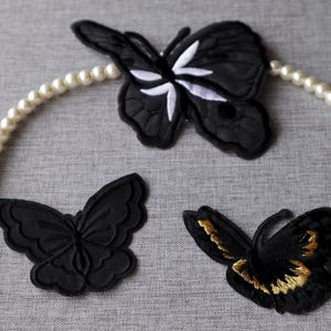 May include: Three black butterfly patches with pearl necklace accents. One butterfly has white details, another has gold accents. The patches are on a grey surface, creating a contrast with the black.