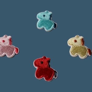 Könnte beinhalten: Vier gestickte Pferdeaufnäher in Rosa, Rot, Gelb und Türkis. Jeder Aufnäher hat eine weiße Umrandung und einen kleinen schwarzen Punkt als Auge. Die Aufnäher sind auf einem blauen Hintergrund angeordnet.