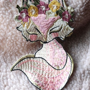 May include: Pink embroidered fox patch with a floral crown of yellow and pink flowers. The fox has a closed-eye expression and a curled tail.