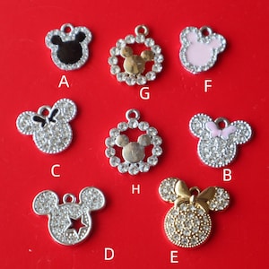May include: A collection of eight metal charms shaped like mouse heads, labeled A through H. The charms feature various designs including black, pink, and gold enamel, clear rhinestones, bows, and star cutouts, each with a small top loop for hanging.