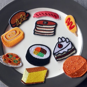 Peut inclure: Divers patchs brodés représentant des aliments, dont un hot-dog, des crêpes, des sushis, un gâteau et un corndog, disposés sur une assiette blanche avec un bord gris. Les patchs présentent des couleurs vives et des coutures détaillées.