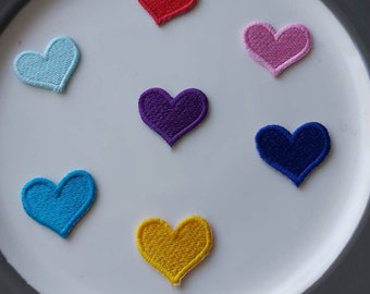 May include: Seven felt heart patches in different colors: red, light blue, pink, purple, blue, yellow, and dark blue. The patches are arranged in a circle on a white plate.