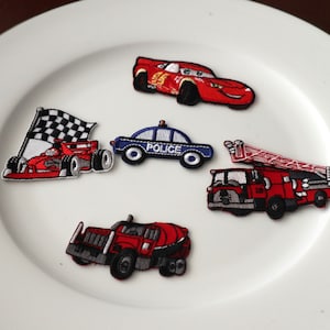 May include: Five embroidered iron-on patches featuring different vehicles. The patches include a red race car with a checkered flag, a blue police car, a red fire truck, a red cement truck, and a red race car with the number 95.