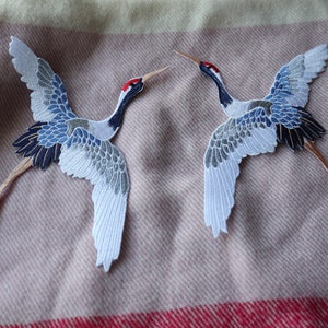 May include: Two embroidered iron-on patches of grey and blue cranes with red beaks and black eyes. The cranes are flying with their wings spread.