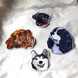 May include: Four embroidered iron-on patches of different dog breeds. The patches are on a white fabric background. The dogs are a black Labrador Retriever, a brown and white Cocker Spaniel, a black and white Cavalier King Charles Spaniel, and a black and white Husky.