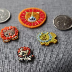 May include: A collection of embroidered lion patches in various designs and colors, including orange, yellow, and brown. The patches are arranged on a gray fabric surface, with two coins visible.