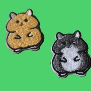 May include: Two embroidered patches of cartoon hamsters. One is tan and the other is black and white. Both have a white border and are set against a green background. The patches are designed with simple, cute details.