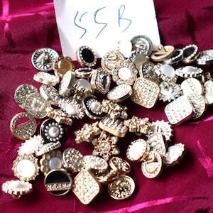 May include: A selection of decorative buttons in diverse shapes and styles, featuring rhinestones, pearls, and metallic finishes. The buttons are scattered on a deep red fabric. The assortment includes round, square, and flower-shaped buttons. A small white tag with the handwritten text "55B" is visible.