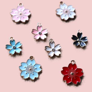 May include: Assortment of eight metal flower charms with enamel paint and rhinestone accents. The charms are in various colors, including pink, white, blue, black, and red.